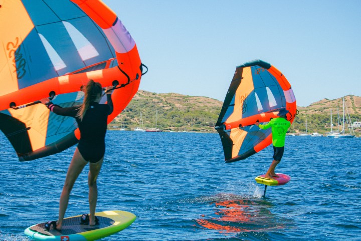 Two people wing foiling on blue water near a hilly coastline.